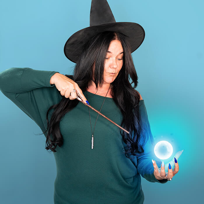 A headshot of a women in a green sweater wearing a witch hat and holding a wand | Catherine Ralls